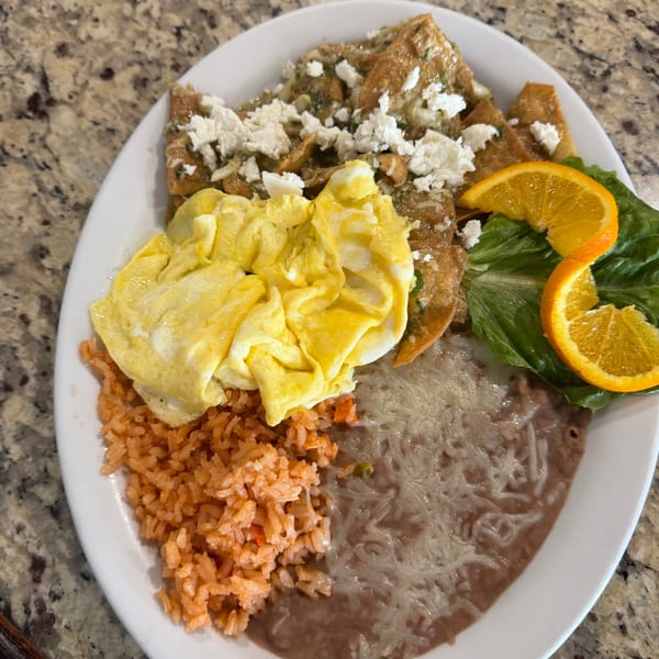 A breakfast plate with scrambled eggs, chilaquiles, rice, refried beans, lettuce, and orange slices.