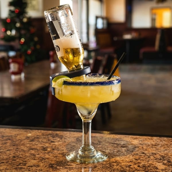 Margarita with an upside-down beer bottle and lime on a bar counter, with a restaurant in the background.