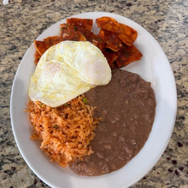 A white plate holds chilaquiles, two fried eggs, Mexican rice, and refried beans on a granite countertop.